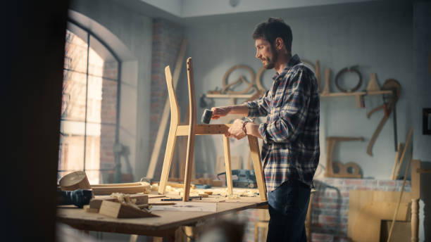 Young Artisan Furniture Designer Marking Out Dimensions on a Blueprint and Starting to Assemble a Wooden Chair. Talented Carpenter Working in a Studio in Loft Space with Tools on the Walls.